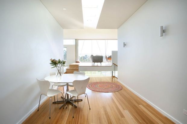 Bright, minimalist open-plan dining and living area with a small round white dining table and sculptural chairs in the foreground. Warm timber flooring extends toward a raised sitting area with a rug and armchair, while full-height glazing brings in abundant natural light. White walls, recessed ceiling lighting and simple wall sconces create a clean contemporary interior.