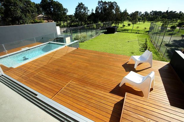 A contemporary outdoor terrace with a broad timber deck, two white sculptural lounge chairs, and an elevated rectangular swimming pool. Glass pool fencing and low privacy walls surround the pool, while a large manicured lawn and fenced recreational courts extend into the landscaped grounds.