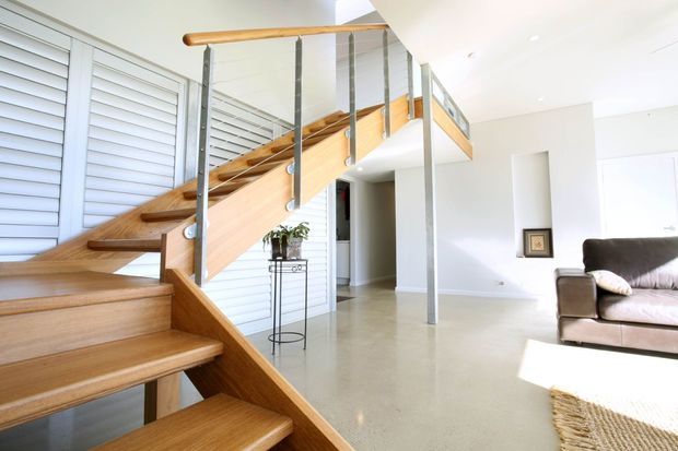 Bright contemporary open-plan interior featuring a sculptural timber staircase with open risers, slim metal balustrade posts, and a timber handrail. White louvred wall panels, polished light flooring, recessed lighting, and a partial leather sofa create an airy living area around the stairwell.