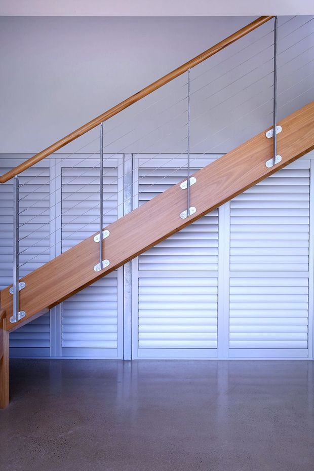 Interior architectural detail showing a modern timber staircase with a warm wood stringer and handrail, slender metal posts, and horizontal cable balustrade. The stair is set against white louvred panels and a pale wall, with a smooth polished concrete floor below.