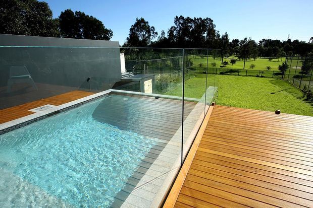 A modern outdoor swimming pool with a shallow tiled section and submerged steps, bordered by expansive timber decking and frameless glass balustrades. A dark rendered wall and nearby modern structure frame the pool, with a broad green landscaped setting beyond.