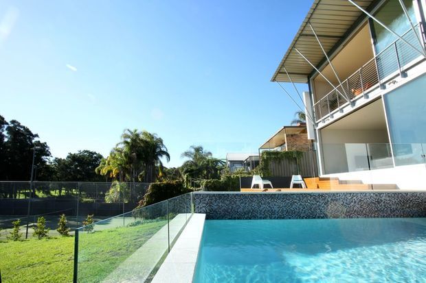 Exterior view of a contemporary multi-level residence beside a swimming pool. The home features light rendered walls, extensive glazing, metal balcony balustrades, a projecting corrugated roof, and a tiled pool edge, with landscaped gardens and neighbouring homes in the background.