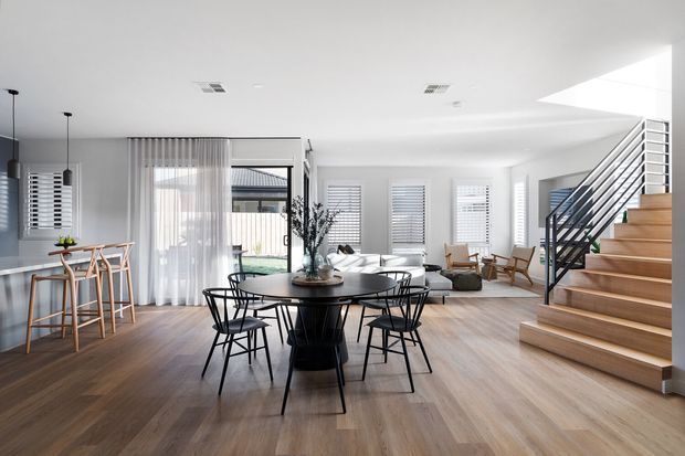 Bright open-plan residential interior combining a dining area, kitchen peninsula, and living room beneath a white ceiling. The room features warm timber flooring, a black round dining table with matching wire-frame chairs, pale upholstered seating, extensive windows with white plantation shutters and sheer curtains, and a timber stair with dark metal balustrade leading to an upper level. A large skylight or clerestory opening brings in strong natural light.