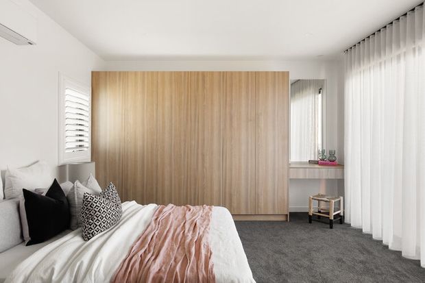 Bright, compact contemporary bedroom with a large wall of warm timber built-in wardrobes, a low upholstered bed with layered white, black and patterned cushions, and a blush-pink throw. Full-height sheer curtains, plantation shutters, grey carpet and white walls create a soft, clean and refined interior.