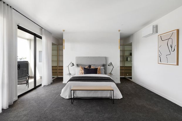 Bright, contemporary bedroom with a centered upholstered bed, dark charcoal carpet, white walls and ceiling, sheer floor-to-ceiling curtains, and black-framed sliding glazing opening to a balcony. Built-in shelving and cabinetry flank the bed, with minimalist bedside lamps, a timber-and-black metal bench, and abstract wall art.
