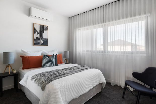 A bright, contemporary bedroom with a neatly made bed dressed in white textured bedding, rust-orange and patterned cushions, and a grey throw. The room includes sheer full-height curtains over a large window, a dark bedside table with a tripod lamp, wall-mounted air conditioning, abstract artwork, and a dark sculptural lounge chair. The palette is predominantly white, grey, black, and warm rust accents.