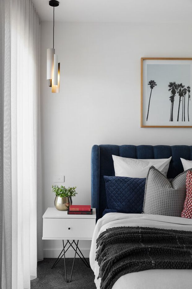 A refined bedroom corner with a deep navy upholstered bed, layered white, grey, navy and patterned cushions, and a dark textured throw. A white floating bedside table with slender black crossed legs sits beside the bed, topped with books and a metallic planter. Floor-to-ceiling sheer curtains provide soft daylight, while a sculptural pendant light hangs beside a framed palm-tree print. The overall design is contemporary, polished and softly layered.