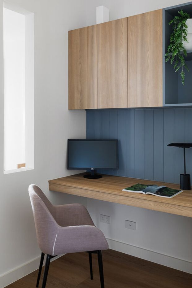 Compact built-in home office nook featuring a wall-mounted timber desk, matching overhead cabinetry, dark blue vertical wall panelling, open shelving with a trailing plant, and a small monitor. A pale upholstered chair sits on dark timber flooring beside a bright window.