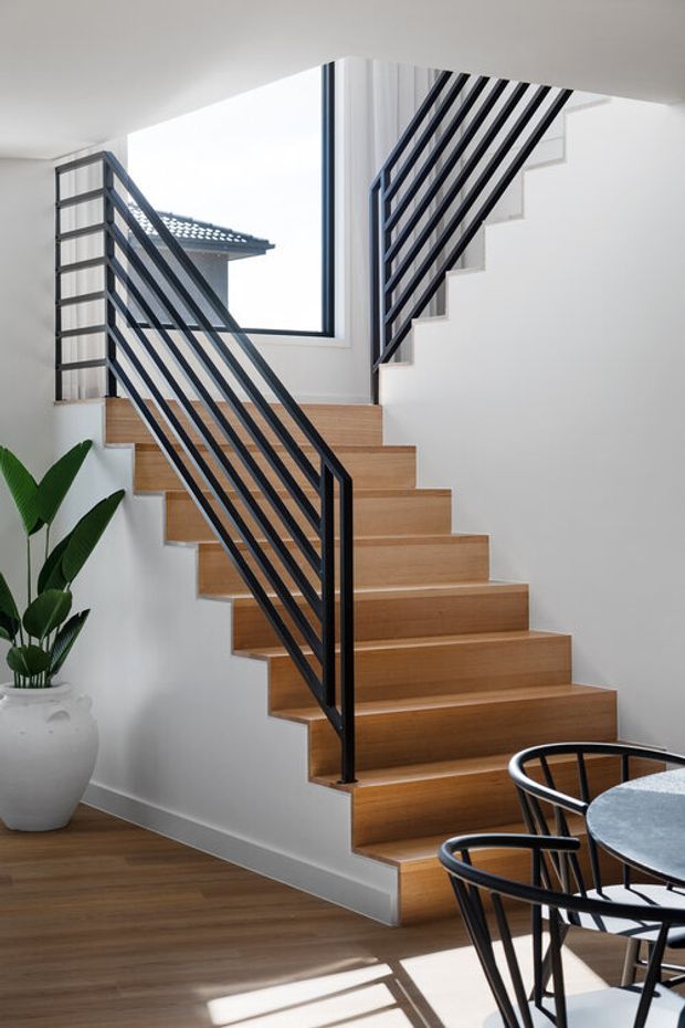 Bright contemporary interior featuring a timber staircase with open risers and black metal balustrades, white walls, a large window, timber flooring, and a small dining area with black chairs in the foreground. A potted plant adds greenery beside the stair.