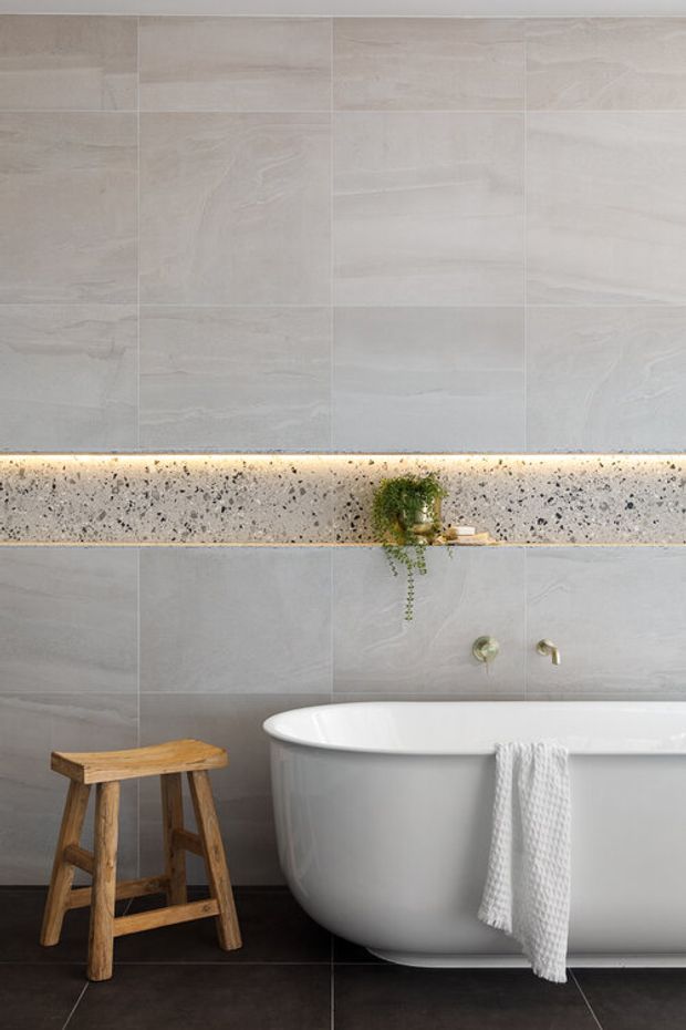A refined bathroom featuring a freestanding white bathtub against large pale grey stone-look wall tiles. A horizontal terrazzo shelf with integrated warm LED lighting spans the wall, holding a small trailing plant and accessories. Dark floor tiles, brushed metallic wall taps, and a rustic timber stool provide contrast.