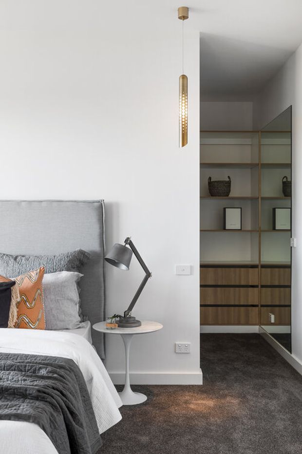 A refined bedroom corner with a grey upholstered bedhead, layered grey and white bedding, and a patterned rust-orange accent cushion. A small white pedestal side table holds a contemporary angled desk lamp. Beside the bed is an open walk-in wardrobe with dark carpet, timber-toned drawers, glass shelving, hanging rails, and integrated storage. A slim brass pendant light hangs in front of the wardrobe. The palette is neutral, soft, and contemporary with warm timber and brass details.