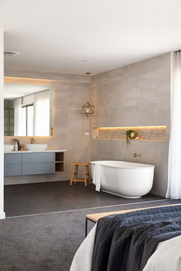 Contemporary open ensuite bathroom viewed from a bedroom, featuring a freestanding white bathtub, wall-mounted blue-grey vanity, large illuminated mirror, warm grey stone-look wall tiles, dark floor tiles, recessed display niche with lighting, and sheer curtains. The foreground shows the edge of the bed, clearly connecting the bathroom to the bedroom.