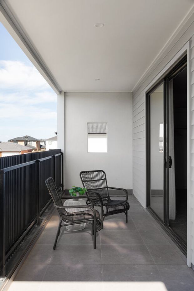 A covered outdoor balcony or terrace with grey tiled flooring, white horizontal cladding, a white ceiling with recessed lights, black-framed sliding glass doors, and dark metal balustrading. Two black wire-frame lounge chairs and a small glass table create a compact sitting area. Neighbouring rooftops are visible beyond the balcony.