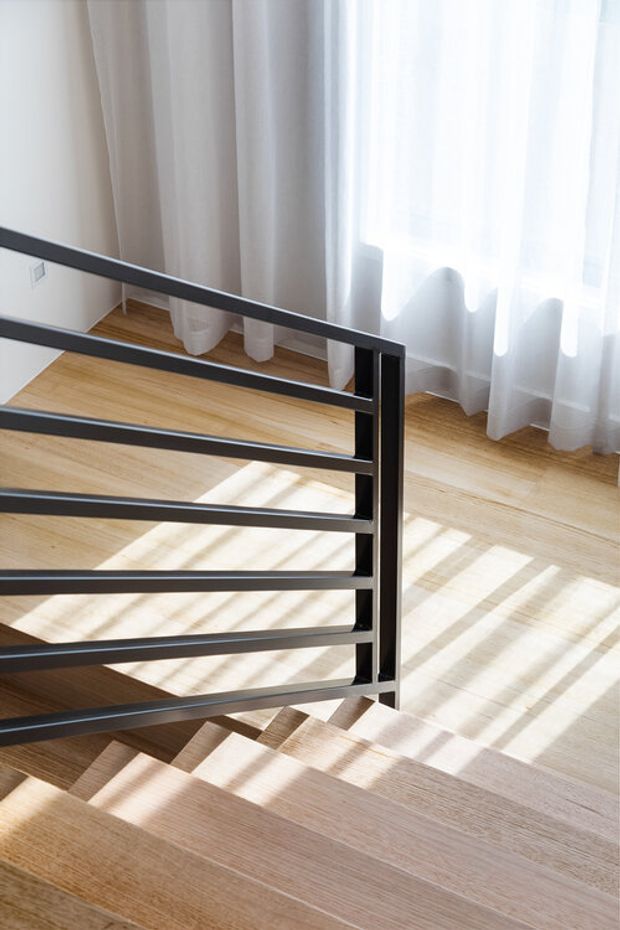 Bright contemporary stairway with pale timber flooring and stairs, a simple black metal horizontal-rail balustrade, and full-height sheer curtains filtering strong natural light.