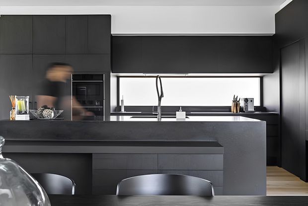 A sleek contemporary kitchen viewed from the dining area, dominated by matte charcoal-black full-height cabinetry and a large dark island with an integrated bench or overhang. The kitchen includes a long horizontal window, stainless or chrome tapware, built-in appliances, and subtle timber flooring extending toward the right. Dark dining chairs and a partially visible dining table occupy the foreground.