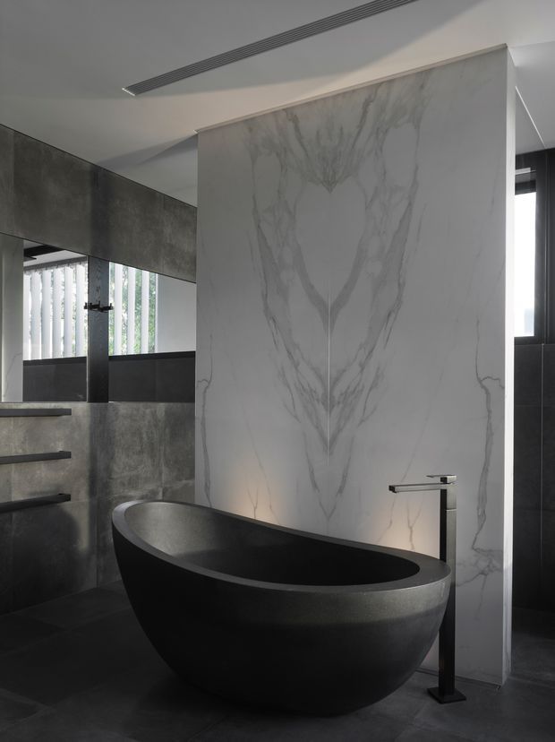 A dramatic contemporary bathroom featuring a freestanding matte black bathtub, dark grey tiled or concrete-look walls and floor, a large white marble-look feature slab with pronounced grey veining, and minimalist black fixtures. A frameless or partially framed shower area with dark finishes is visible to the left, while vertical blinds and a window provide filtered natural light.