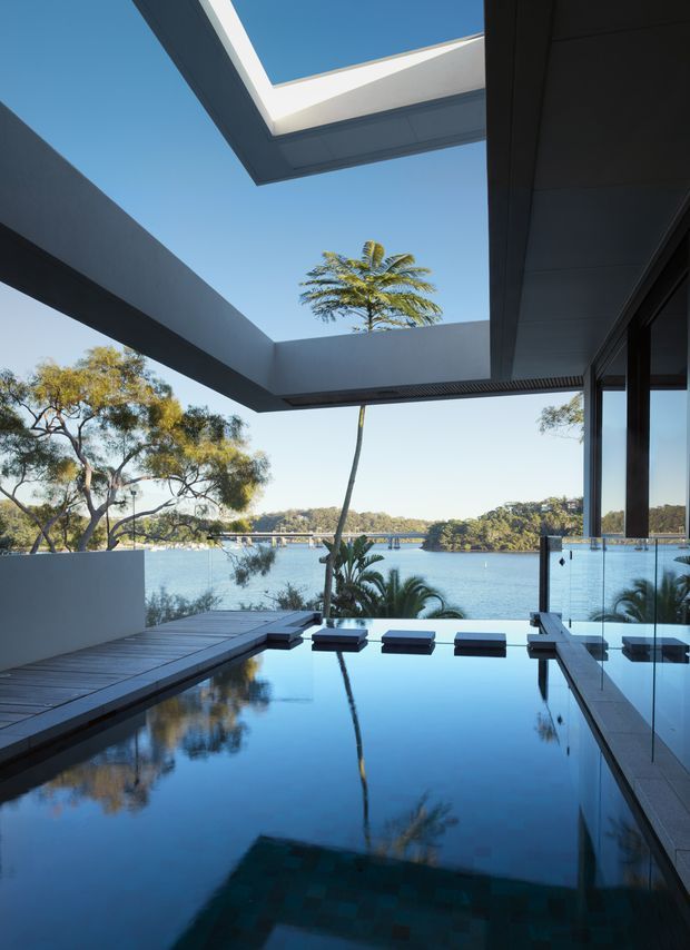 A contemporary waterfront architectural view from a covered outdoor terrace, featuring a reflective infinity-edge swimming pool, dark timber decking, broad cantilevered roof planes, and full-height glazing. A tropical palm and established trees frame the calm blue water beyond, while a large overhead opening creates a dramatic sky-lit courtyard effect.