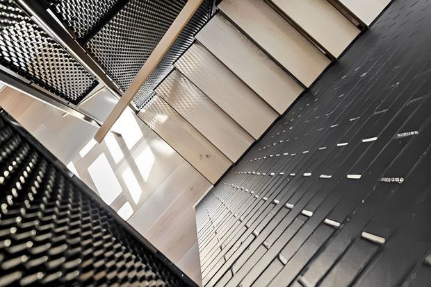 Upward-looking architectural interior view of a dramatic stairway or landing. Pale timber stair treads and a slim timber handrail contrast with dark perforated metal or mesh balustrades and a dark textured wall. Strong daylight enters through a skylight or clerestory opening, creating geometric light and shadow patterns. The image emphasizes the building's circulation space, material contrast, and sculptural detailing.