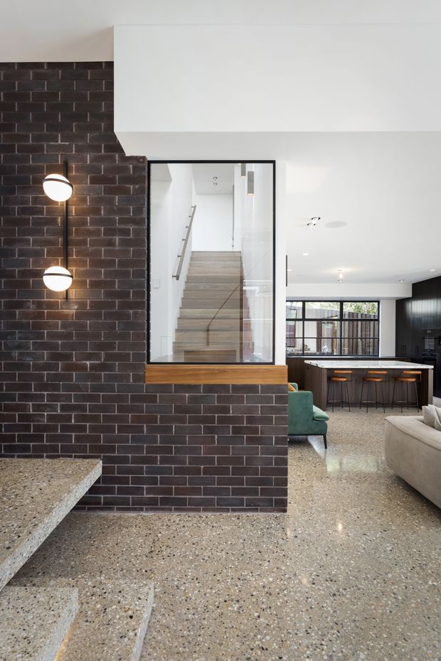 A contemporary open-plan interior featuring a dark charcoal brick partition wall with a large black-framed internal window overlooking a stairwell. The foreground has polished terrazzo flooring and a terrazzo stair edge, while the background opens to a kitchen with a dark island, timber bar stools, and black-framed glazing. A neutral sofa and green armchair are partially visible, with spherical wall lights mounted on the brickwork.
