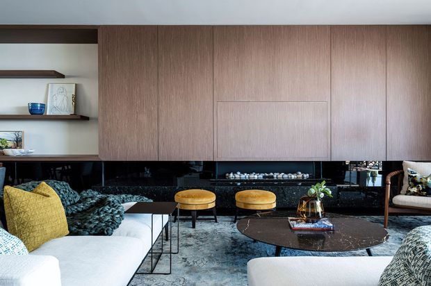 Refined contemporary living room featuring extensive warm wood built-in cabinetry and shelving, a dark glossy stone or marble fireplace wall, pale sofas, patterned textiles, rounded ottomans, and a low sculptural coffee table. The room has a sophisticated, layered palette with strong horizontal cabinetry and built-in display storage.