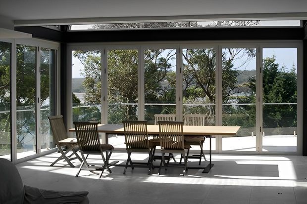 Bright enclosed dining area with a long rectangular timber dining table and folding timber chairs, surrounded by extensive floor-to-ceiling glazing and glass doors. The room opens visually to a leafy outdoor setting and has a dark structural ceiling frame with a linear skylight or clerestory strip.