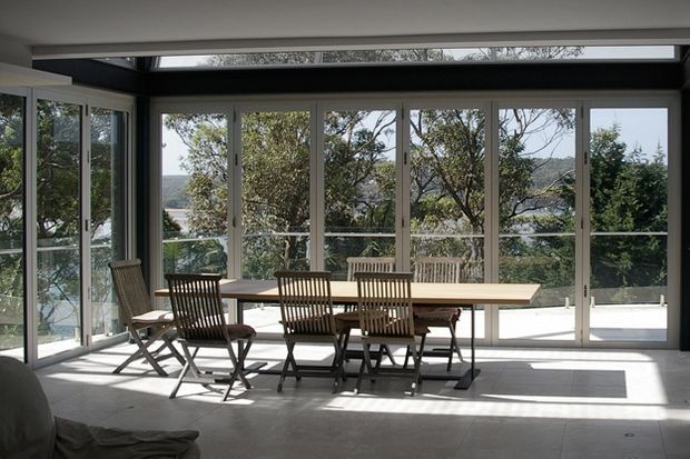 Bright dining area with a long timber dining table and matching chairs positioned beside extensive floor-to-ceiling glazing. The space opens onto a balcony or terrace and overlooks a leafy, hilly landscape, with strong natural light, pale flooring, and dark framing details.