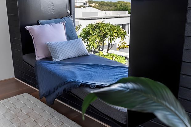 A compact bedroom nook with a single built-in bed positioned beside large glazing, dressed in deep blue bedding and pastel blue and lilac cushions. Dark brick or tiled wall surfaces frame the bed, with timber flooring and a pale patterned rug. A leafy indoor plant partially obscures the foreground, while neighbouring buildings and greenery are visible outside.