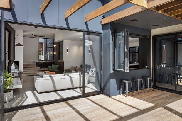 A bright contemporary indoor-outdoor entertaining area with a timber-lined covered terrace opening through large black-framed glazed doors into a living room. The interior includes white sofas, a dark kitchen or bar zone with stools, polished concrete flooring, exposed timber beams, and high-level glazing.