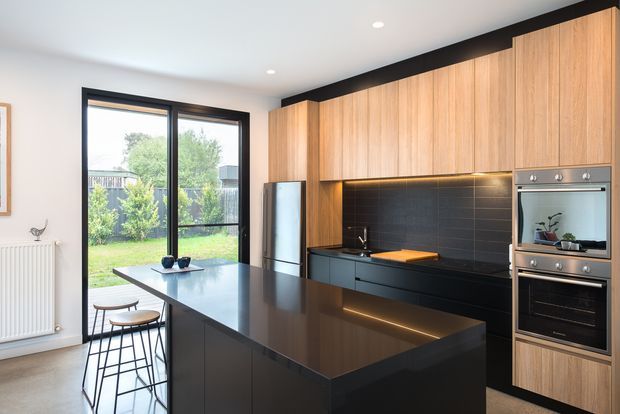 A bright contemporary kitchen with a large black island and integrated breakfast seating, full-height warm timber cabinetry, dark tiled splashback, black benchtops, and stacked stainless-steel ovens. Floor-to-ceiling glazing opens to a garden, while subtle under-cabinet lighting adds warmth.