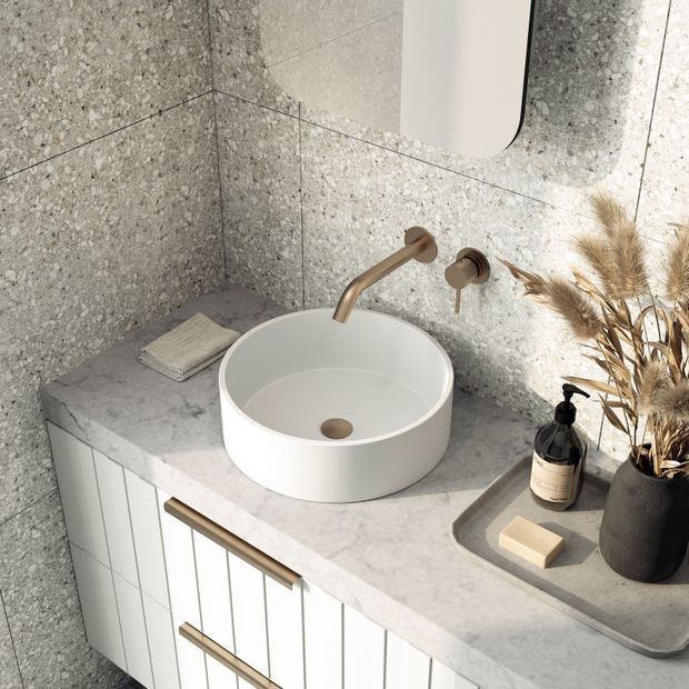A close-up view of a refined bathroom vanity featuring a round white vessel basin, wall-mounted brushed brass tapware, a pale stone benchtop and vertically fluted white cabinetry. Grey terrazzo-style wall tiles continue behind the vanity, with a rounded-corner mirror, soap dispenser, tray and dried grasses adding understated styling.