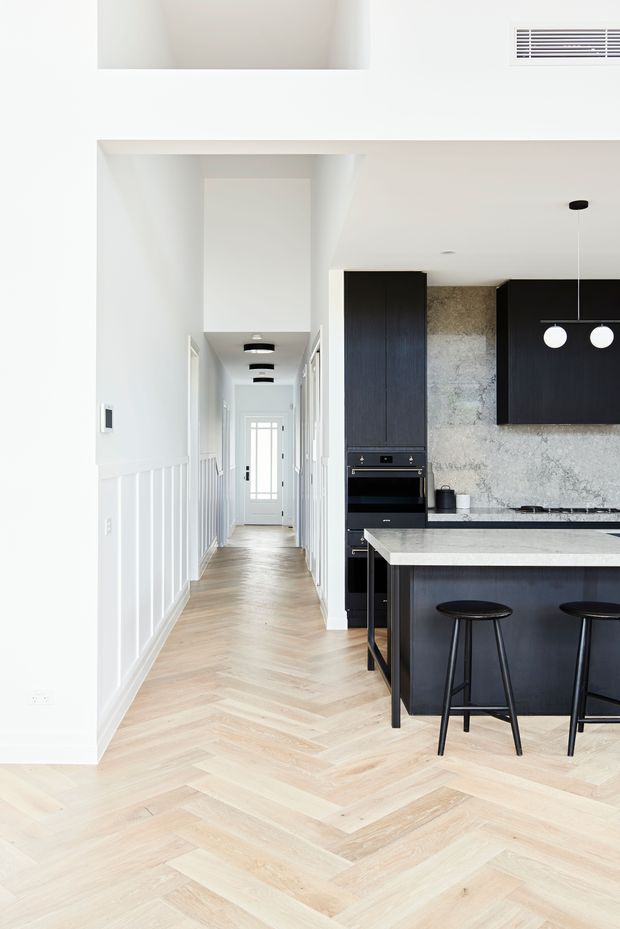 Bright contemporary open-plan kitchen viewed from an adjoining hallway, featuring a black island with stools, dark built-in cabinetry, pale stone benchtops and splashback, integrated appliances, white wall panelling, and light timber herringbone flooring. A long hallway with recessed and pendant lighting leads to a glazed entry door.