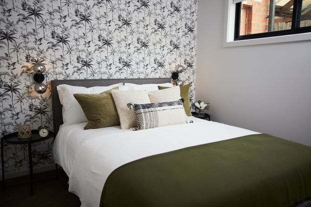 A styled bedroom with a double bed dressed in white bedding and an olive-green throw, layered pillows, and a dark timber headboard. One feature wall has monochrome botanical wallpaper, while the remaining walls are painted grey. Black bedside tables, globe wall lights, and a small window contribute to the contemporary, botanical-inspired scheme.