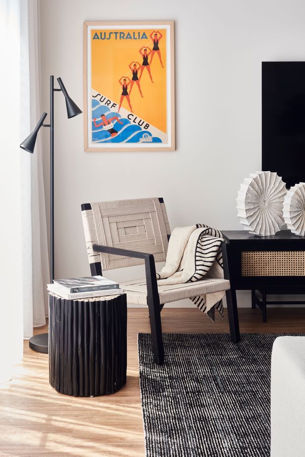 A stylish living room corner with a warm neutral palette, light timber flooring, a black-and-white textured rug, a woven lounge chair draped with a striped throw, a black fluted side table, and a dark media console with cane-front detailing. A black floor lamp, framed vintage Australia surf poster, sculptural paper wall decor, and partial television add graphic and artistic accents. Natural light filters through sheer curtains.