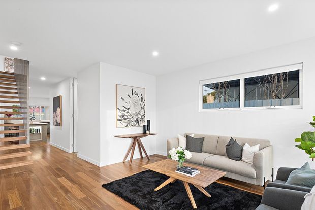 Bright contemporary living room with white walls and recessed ceiling lights, warm timber flooring, a beige sofa and grey armchairs arranged around a low timber coffee table on a black rug. A minimalist console table with artwork sits against the wall, while an open timber staircase and hallway are visible to the left.