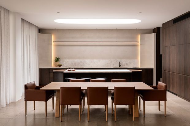 A refined open-plan dining and kitchen interior. A long timber dining table with six brown leather chairs faces a dark timber kitchen island and a full-height textured stone or plaster splashback with integrated shelving and warm concealed lighting. Floor-to-ceiling sheer curtains line the left side, while dark timber cabinetry occupies the right wall. A large oval recessed ceiling light provides a soft contemporary focal point.