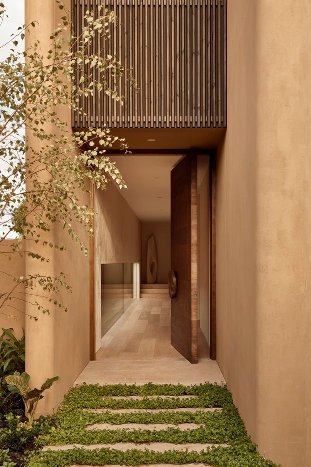 A warmly toned residential entrance viewed from an exterior garden walkway. Pale earthy plaster walls frame a recessed entry with broad steps edged by low greenery. A large dark timber pivot door is open to a long interior hallway, while a vertical timber slatted screen projects above the entrance. The hallway continues toward a reflective glazed or mirrored surface and features timber-toned flooring and subtle wall artwork. The composition is calm, tactile, and contemporary, with strong vertical lines and a sheltered threshold.