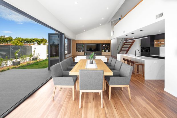 Bright open-plan contemporary living, dining and kitchen interior with a large timber dining table surrounded by upholstered chairs, built-in timber media cabinetry, white kitchen cabinetry and island, and a timber staircase leading to an upper level. Full-height sliding glazing opens onto a paved outdoor area and landscaped garden. The room has a high ceiling and abundant natural light.
