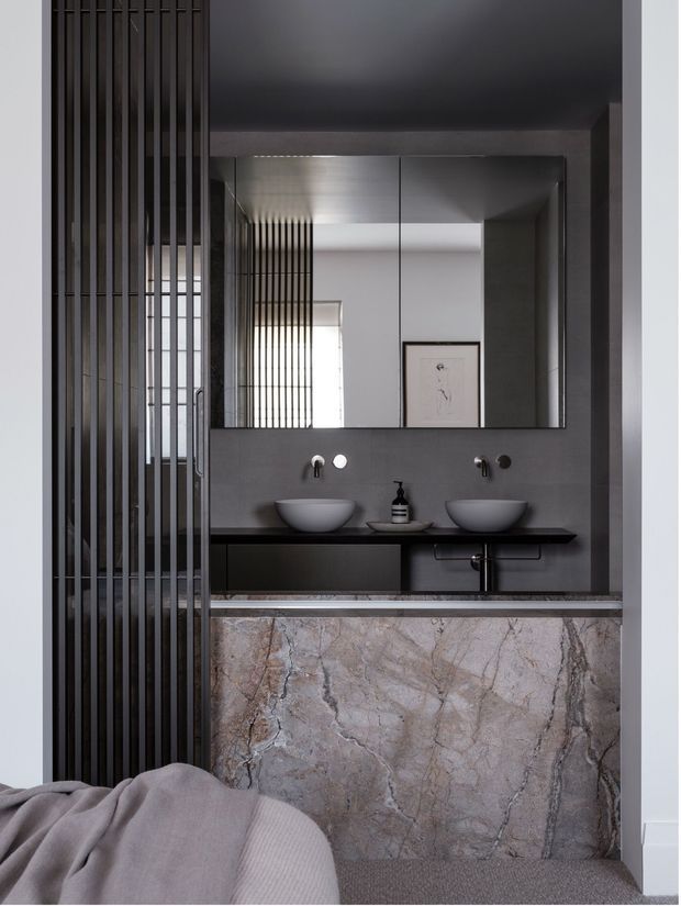 A refined dark-toned bathroom vanity viewed through a vertical black metal screen, with twin vessel basins, wall-mounted taps, a large frameless mirror, and a dramatic veined stone-clad vanity front. A bedroom with bedding is visible in the foreground, establishing a direct connection between the bathroom and bedroom.