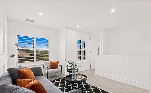 Bright contemporary living room with white walls, recessed ceiling lights, plantation shutters, grey sectional sofa, orange cushions, black metal-framed occasional chairs, round black coffee tables, and a black-and-white patterned rug. A half-height wall or stair balustrade defines the right side of the room, with greenery visible through the windows.