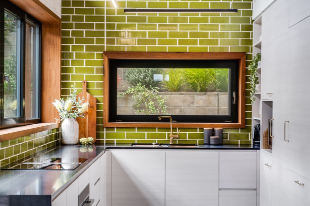 A compact contemporary kitchen with a bold lime-green glazed brick-tile backsplash, white flat-panel cabinetry, dark stone benchtops, and warm timber-framed windows. The space includes a cooktop, sink beneath a timber-framed window, integrated storage, and black and brass-toned fixtures. Natural light enters through the windows, with greenery visible outside.