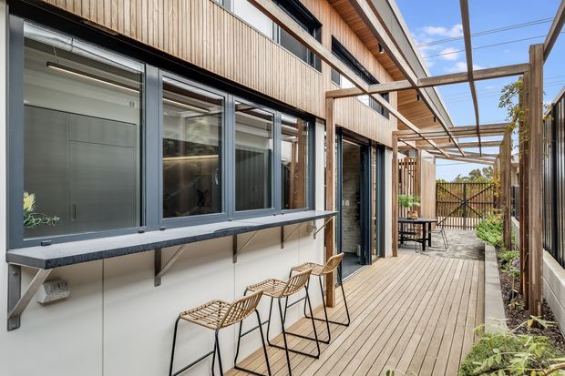 A contemporary timber-clad residential exterior with a long timber deck and covered outdoor walkway. Floor-to-ceiling dark-framed glazing opens onto the deck, with a narrow built-in counter and bar stools along the facade, plus a small outdoor dining setting. A timber pergola, vertical privacy fencing, and landscaped planting define the narrow courtyard-like outdoor space.