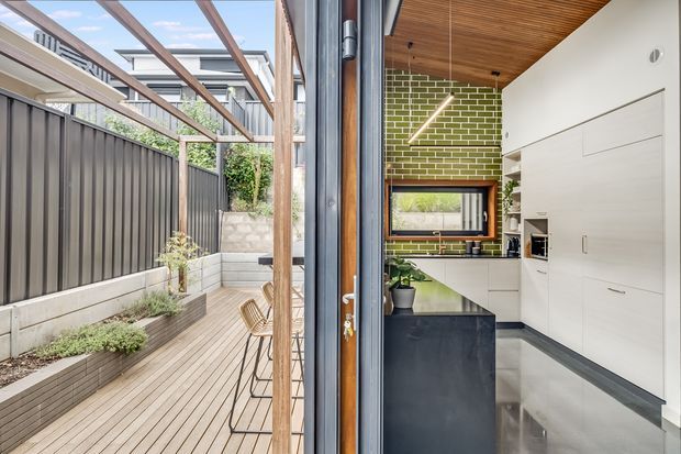 View through an open glazed door from a compact contemporary kitchen to a narrow timber deck courtyard. The kitchen features white built-in cabinetry, a dark island, polished concrete flooring, a green glazed brick splashback, timber-lined ceiling, and a warm timber-framed window. Outside, the deck is bordered by a dark vertical metal fence, raised planter beds, and a simple timber pergola.