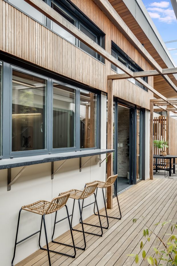 A contemporary multi-level building facade viewed from a timber deck or terrace. The exterior combines vertical natural timber cladding, dark aluminium-framed glazing, exposed concrete bands, and timber soffits. A long outdoor bar counter with woven bar stools runs along the lower facade, while a pergola-like timber structure extends over the deck. An open glazed doorway reveals a partially visible interior with shelving and plants.