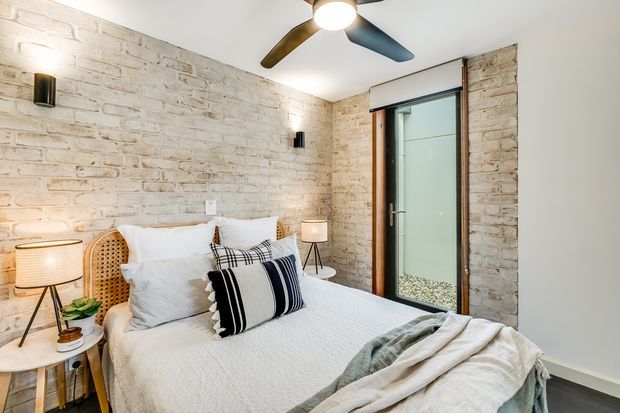A bright, cozy bedroom with a neatly made bed, white bedding, striped accent cushions, and a soft throw. The room features exposed whitewashed brick walls, a timber-framed frosted-glass door, warm bedside lamps, woven rattan details, and a contemporary three-blade ceiling fan.