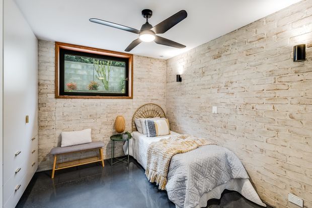 A compact bedroom with exposed white-painted brick walls, a single bed, woven rattan headboard, light bedding, a small bench, and a side table. The room has a dark polished concrete floor, a timber-framed window, recessed storage cabinetry, and a contemporary ceiling fan with integrated light.