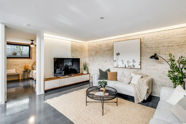 Bright contemporary living room with a white-painted exposed brick feature wall, polished grey concrete flooring, a low timber-and-white media console, neutral sofas, woven textiles, a round black metal coffee table, and integrated perimeter lighting. An adjoining room is visible through a wide opening on the left.