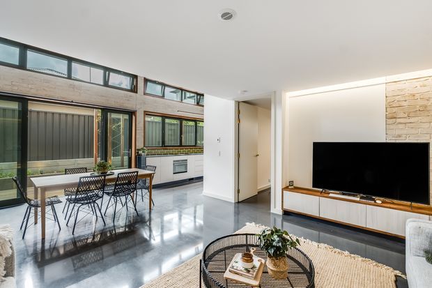 Bright open-plan interior combining a dining area, compact kitchen and living room. The space features polished grey concrete flooring, large industrial-style black-framed glazing, white walls, a timber-accented TV unit, and a white brick feature wall. A dining table sits beside the kitchen, while a round coffee table and sofas define the living area.