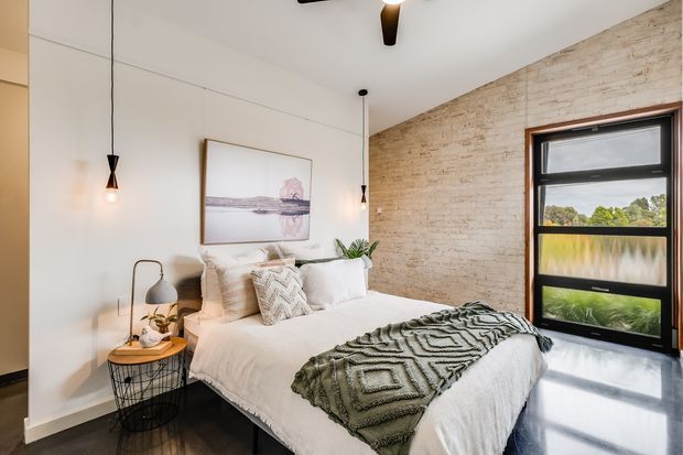 A bright, compact bedroom with a white bed dressed in layered neutral bedding and a textured green-grey throw. The room combines a white painted wall with an exposed pale brick feature wall, dark polished flooring, black-framed glazing, suspended pendant lights, a small bedside table, and understated artwork and plants. The overall feel is warm, refined, and contemporary with industrial accents.