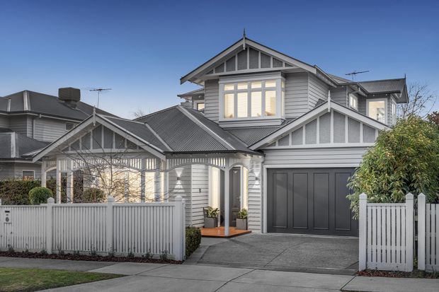 Front exterior view of a large multi-level home with grey horizontal weatherboard cladding, multiple gabled roof sections, decorative white gable detailing, a covered entry porch, upper-level bay window, and an attached double garage. The property is enclosed by a white picket-style fence and landscaped with shrubs.