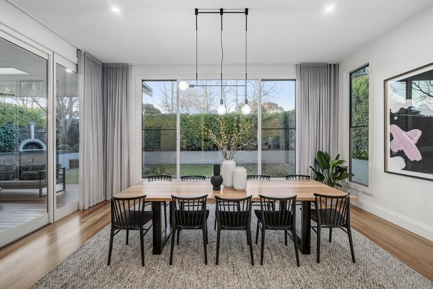 Bright contemporary dining room with a long light-wood dining table surrounded by black spindle-back chairs, set beneath three pendant lights. Full-height glazing and sliding doors open to a leafy garden, with sheer grey curtains, timber flooring, a large neutral rug, indoor plants, and framed artwork. A reflected living area is visible through the glazing.